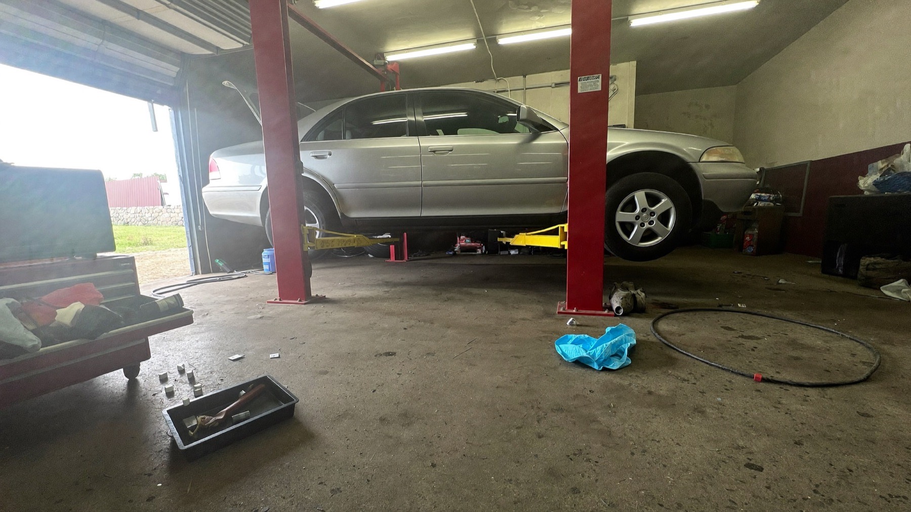 A sedan raised on a two-post lift inside the service bay, bay door open to daylight.